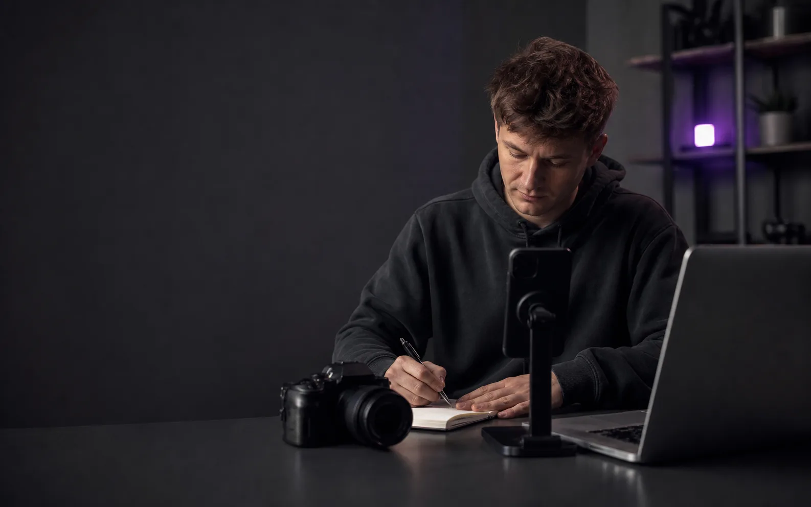 A creator preparing a video at a desk with a camera, phone, notebook, and laptop